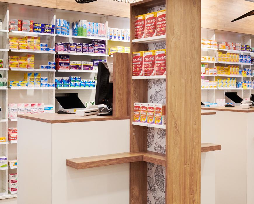Détail d'un poste d'accueil pharmaceutique avec colonne de rangement en chêne clair, tablette PMR et éclairage soigné, mobilier sur mesure pour officine.  