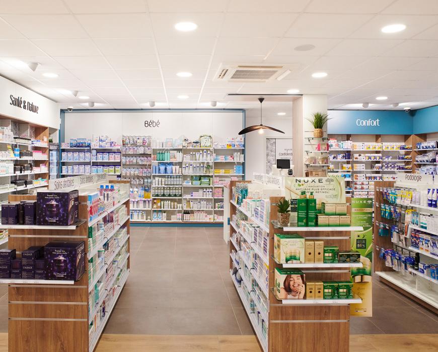 Vue d'ensemble de l'espace de vente d'une pharmacie de 200m², secteurs soins et bébé, sol imitation parquet et plafond lumineux, par JBCC Agenceur