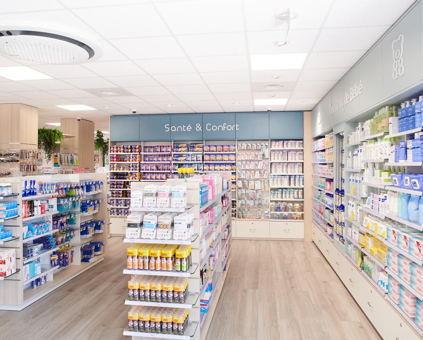 Allée centrale de pharmacie structurée par univers avec gondoles basses et signalétique « Santé & Confort ».